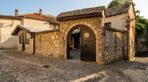 A semicircular stone arched gate with massive wooden doors leading into the cobbled courtyard of a historic building in Mostar