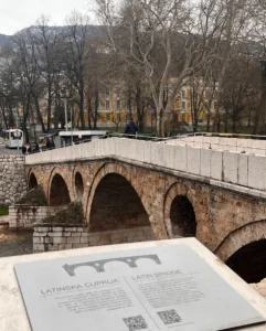Latinska ćuprija u Sarajevu zimi sa info tablom u prvom planu, kameni most bez lišća na drveću