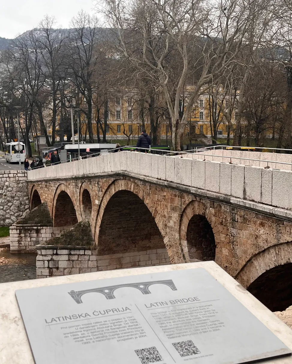 Latinska ćuprija u Sarajevu zimi sa info tablom u prvom planu, kameni most bez lišća na drveću