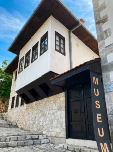 The stone facade and traditional white doksat of the Džemal Bijedić Memorial House, a department of the Museum of Herzegovina, with the vertical "MUSEUM" sign prominently displayed on the black entrance doors