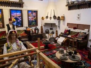 A rich ethnographic display showing a traditional interior with a weaving loom, a doll in folk costume, colorful woven rugs, and old dishes hanging above the hearth