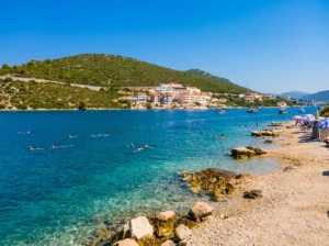 View of the pebbly shore of Jazina Beach in Neum with clean turquoise water where swimmers are bathing, while a green hill and coastal buildings can be seen in the background under a clear sky