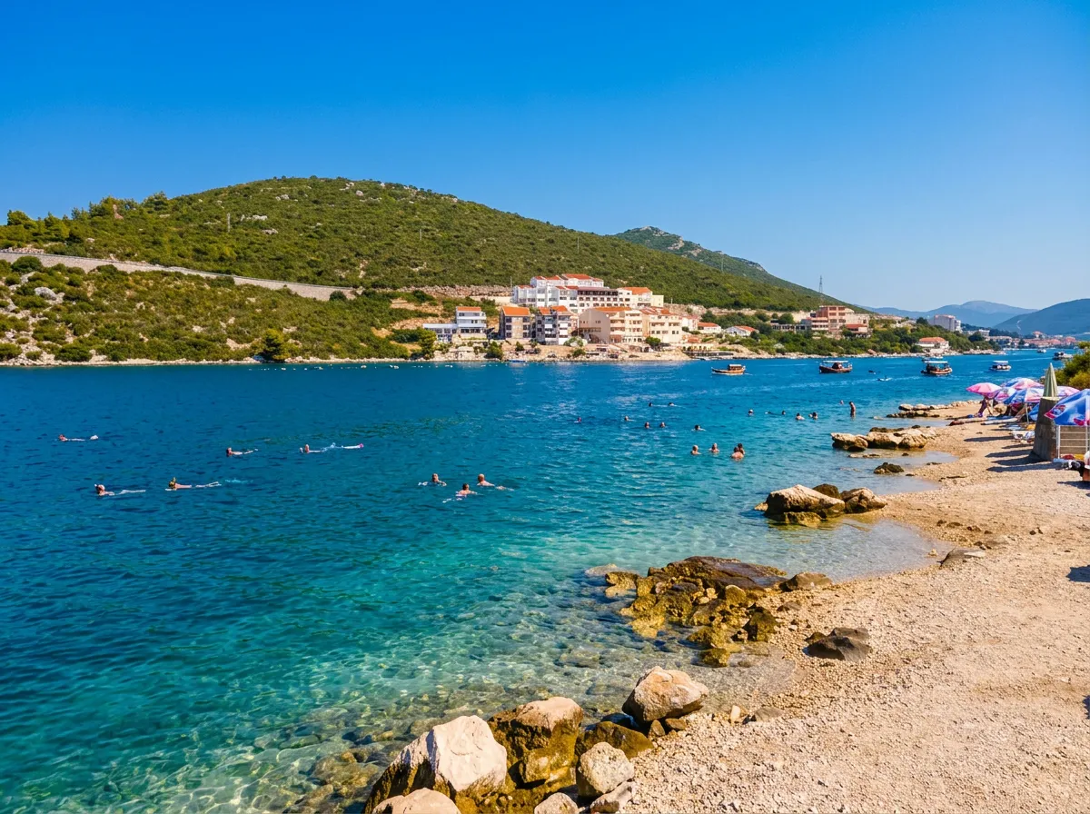 View of the pebbly shore of Jazina Beach in Neum with clean turquoise water where swimmers are bathing, while a green hill and coastal buildings can be seen in the background under a clear sky