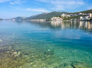 Crystal-clear sea surface with a clearly visible stone seabed in the shallows, with a panorama of Neum and mountain ranges in the distance 