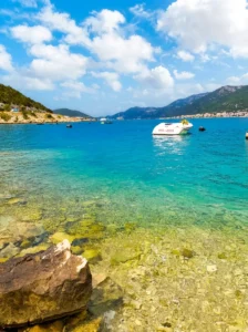 A vivid view of the turquoise-blue bay in Neum with a stone shoreline in the foreground, boats in the distance, and a wide sunny sky with white clouds