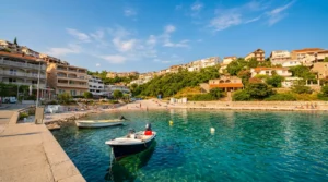 Aerial view of a calm, clear green bay with two small boats moored in front of a pebbly beach, surrounded by apartments and Mediterranean greenery