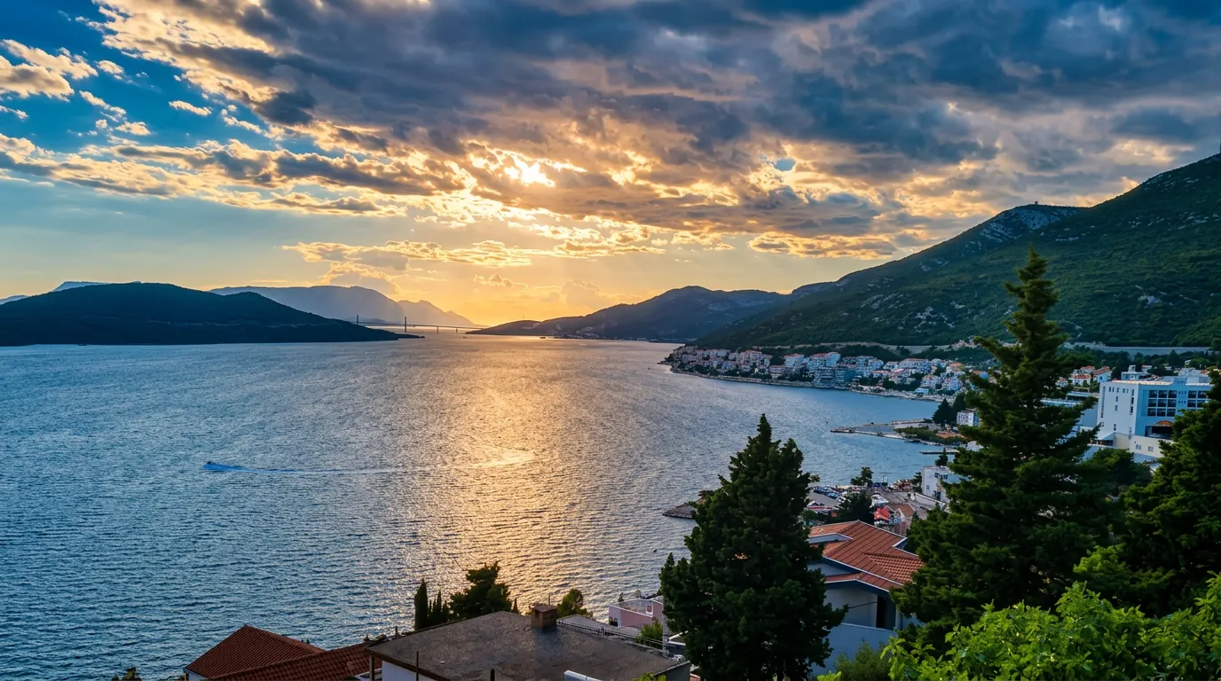 A beautiful panoramic view of Neum Bay at dusk, with dramatic clouds through which golden rays of the setting sun break through and illuminate the sea surface and the coastal rooftops of houses