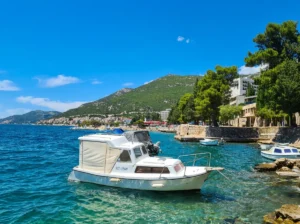 A white motorboat with a canopy anchored on calm turquoise water, while a crowded beach with swimmers, hotels, and a tall green hill under a clear sky can be seen in the background