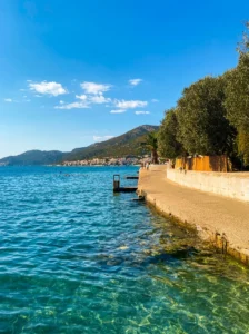 A concrete seaside promenade lined with trees and palm trees, overlooking the shallow, crystal-clear sea with a visible seabed and the distant hills of Neum Bay
