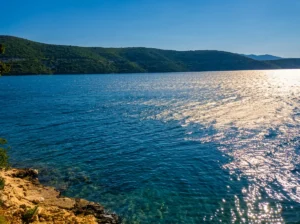 Close-up of shimmering, clear blue sea water in Neum, photographed from the rocky shore and Mimoza Beach in town, with a hilly peninsula in the background under the sun