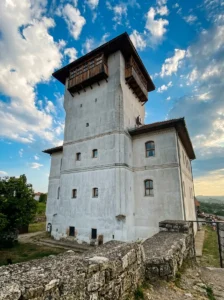 Blick von unten auf den dreistöckigen Weißen Turm in Gradačac mit dem markanten hölzernen Čardak an der Spitze, unter blauem Himmel und leichten Wolken