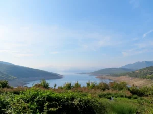 A wide panoramic view from a hill over the blue expanse of Bilećko jezero surrounded by rugged Herzegovinian hills, with green shrubs in the foreground under a bright sky