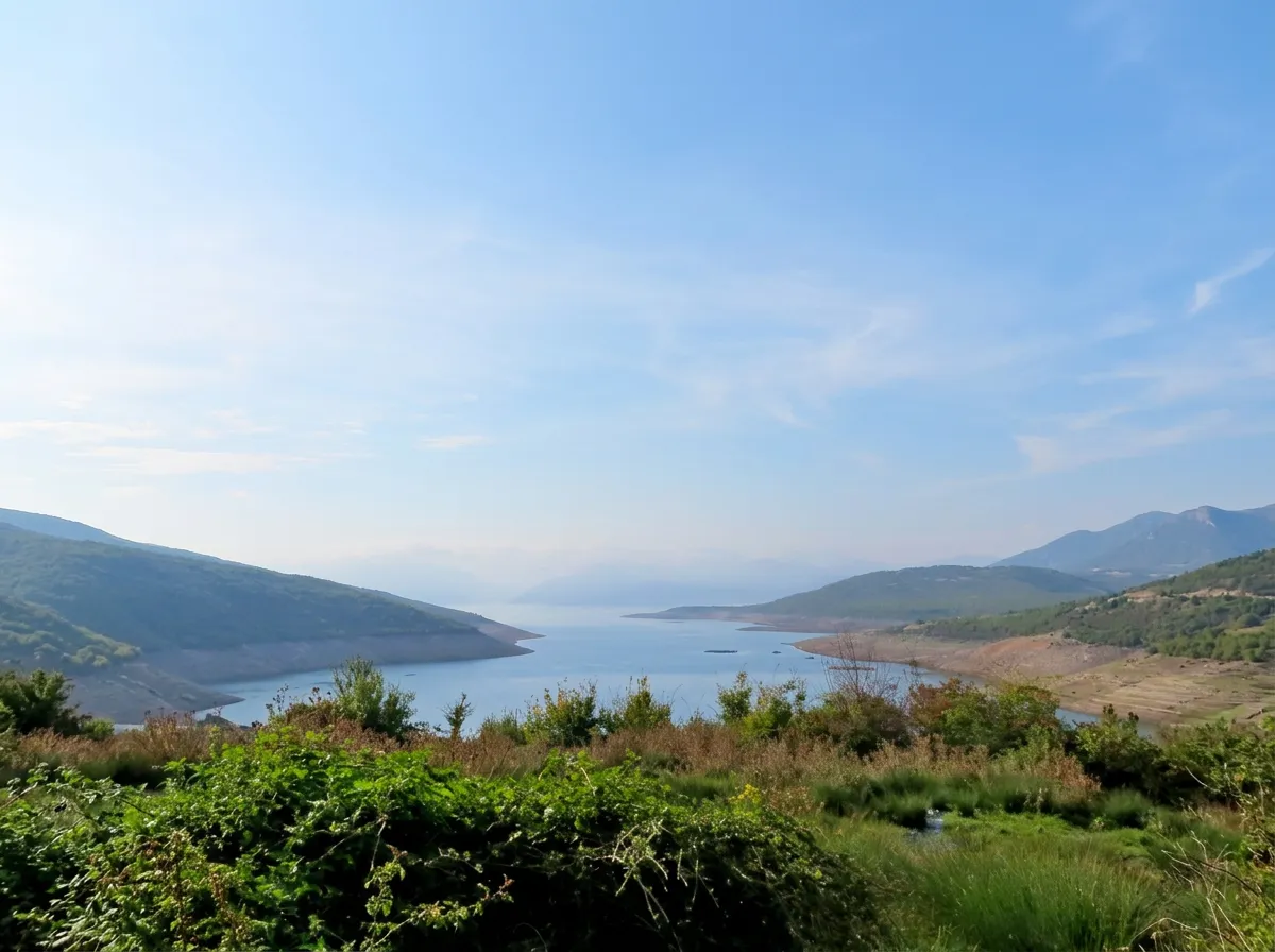 A wide panoramic view from a hill over the blue expanse of Bilećko jezero surrounded by rugged Herzegovinian hills, with green shrubs in the foreground under a bright sky