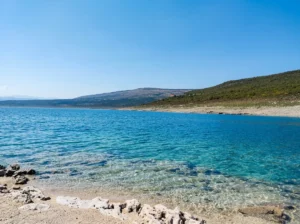 A view from a shallow rocky beach of the beautiful, crystal-clear and transparent turquoise water of Bilećko jezero, which turns dark blue in the distance under a clear sky