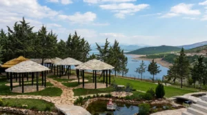 A complex of traditional wooden summer houses and canopies with stone roofs on a landscaped green lawn with paved paths, overlooking pine trees and a lake in the background