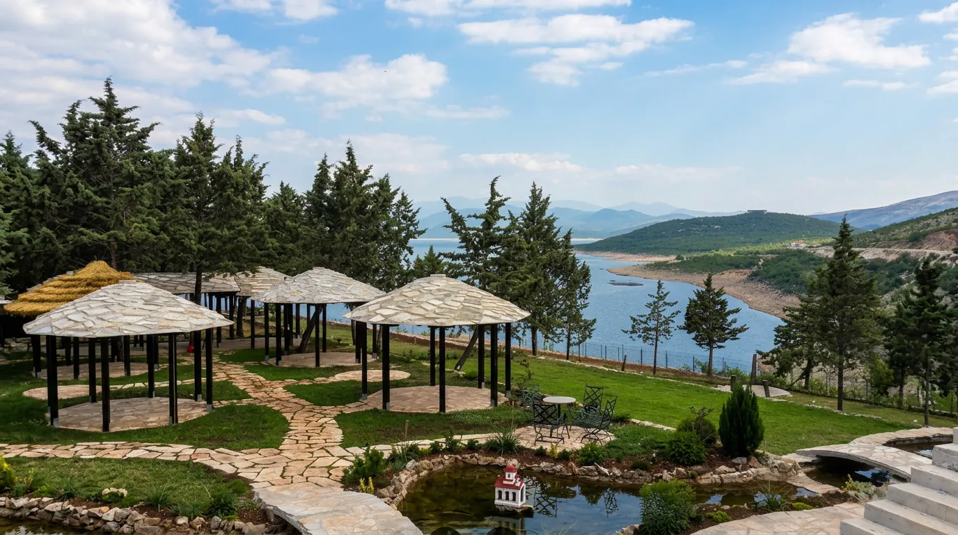 A complex of traditional wooden summer houses and canopies with stone roofs on a landscaped green lawn with paved paths, overlooking pine trees and a lake in the background