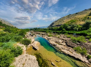 A wide, layered rocky riverbed with pools of clear emerald water, green shrubs along the bank, and hills under a blue sky in the background
