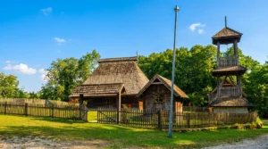 Pogled sa udaljenosti na istorijsku crkvu brvnaru ograđenu drvenom tarabom, okruženu travnatim dvorištem i gustom zelenom šumom pod vedrim nebom