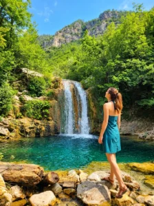 Ein Mädchen in einem blauen Sommerkleid steht am steinigen Ufer und betrachtet den wunderschönen Wasserfall Crni vir, der in ein klares, türkisfarbenes Naturbecken stürzt, umgeben von dichtem Wald