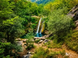 Blick durch üppiges grünes Dickicht und wilde Natur auf den schmalen, hohen Wasserfall Crni vir, der in einen tiefen und ruhigen smaragdgrünen Flusswirbel am Fuße des Hügels stürzt