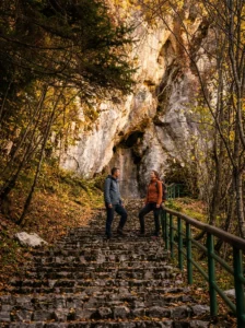 Two hikers stand and talk on a stone staircase with a protective wooden railing that leads through the autumn forest toward the large rocky opening of Djevojačka pećina