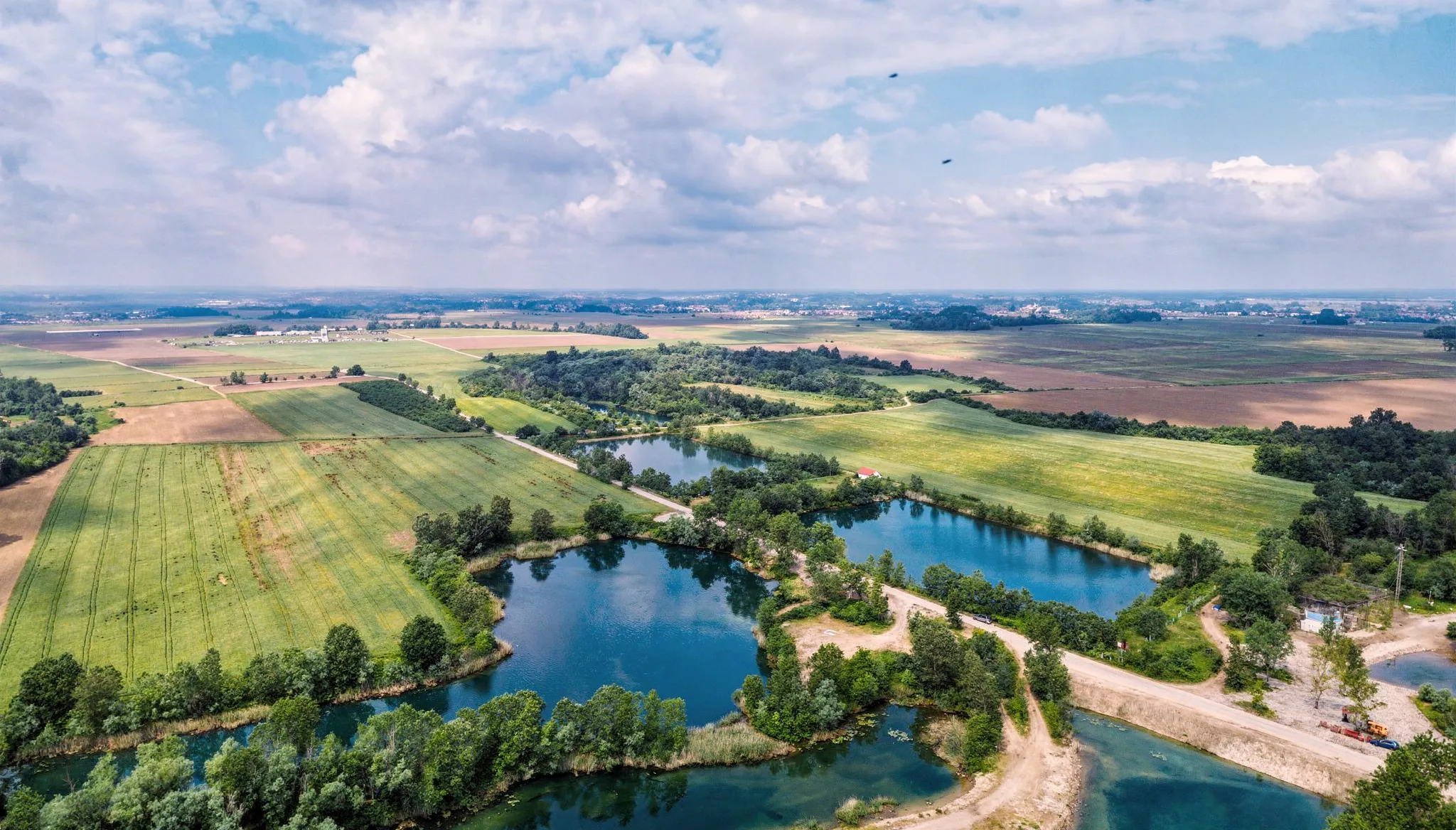 A panoramic bird's-eye shot showing several beautiful blue lakes surrounded by green fields, trees, and gravel paths under a wide, cloudy sky