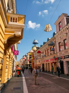 A view along the promenade in Gospodska ulica with passersby above whom creative decorations in the shape of colorful lampshades hang, framed by the architecture of old buildings under a blue sky