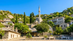 The tall stone minaret and main dome of Hadži-Alijina džamija bathed in sunlight, framed by slender cypress trees and greenery under a clear blue sky