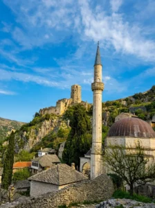 A view of the minaret and dome of Hadži-Alijina džamija, while in the background the historic medieval tower of Gavrankapetanovića rises on a steep hill in Počitelj