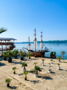 A large wooden pirate ship with masts and unfurled white sails anchored beside a landscaped concrete shore decorated with potted palms, with the calm lake water in the background