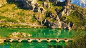 Krupni plan starog lučnog mosta iznad intenzivno tirkizno-zelene površine jezera Grabovica, sa krševitim planinskim padinama u pozadini koje obasjava sunce