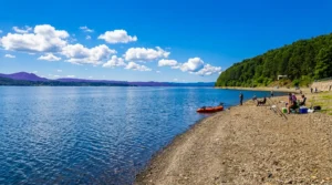 A view along an empty pebble beach toward the calm, dark blue surface of Lake Modrac, with a red boat in the shallows and a wooded hill on the right under a blue sky with white clouds
