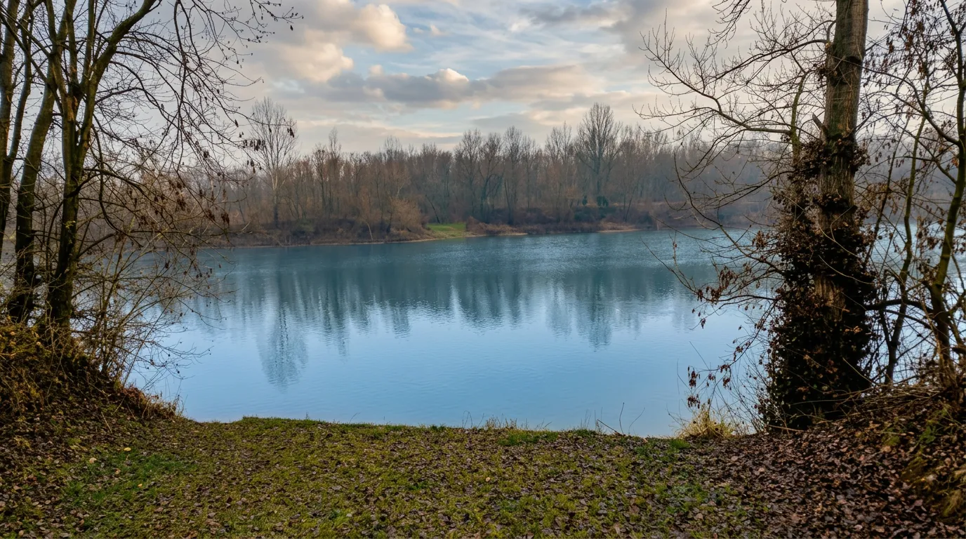 Pogled sa travnate obale na plavičastu površinu jezera Pelagićevo i šumu na suprotnoj strani pod slojevitim oblačnim nebom u prohladan dan