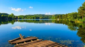 A damaged wooden pier or raft in the foreground on the calm surface of Vidara Lake, reflecting the blue sky and wooded hills in the background