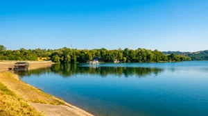 A view over the concrete shore to the clear turquoise-blue water of the lake, where dense trees on the opposite side are clearly reflected under the open sky