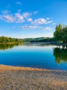 A view from a sunny pebbled shore of the calm and clear blue surface of Vidara Lake, surrounded by green trees and hills under a blue sky with white clouds