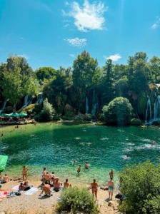 Eine große Zahl von Badegästen am Ufer und im klaren, smaragdgrünen Wasser des natürlichen Beckens vor den prächtigen, von dichtem Grün umgebenen Kravica-Wasserfällen