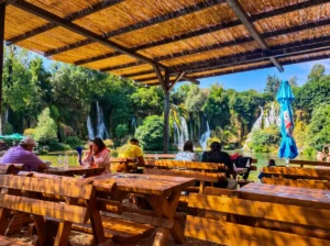 Blick von der sonnigen Holzterrasse des Restaurants unter einem Strohdach auf die Gäste, die an den Tischen sitzen und den Blick auf die Wasserfälle im Hintergrund genießen