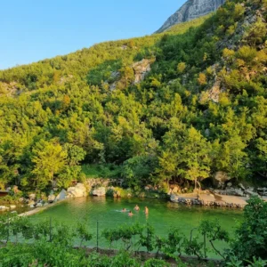 Blick von oben auf ein smaragdgrünes Flussbecken, in dem sich mehrere Badegäste befinden, am Fuße eines steilen, dichten grünen Hügels gelegen