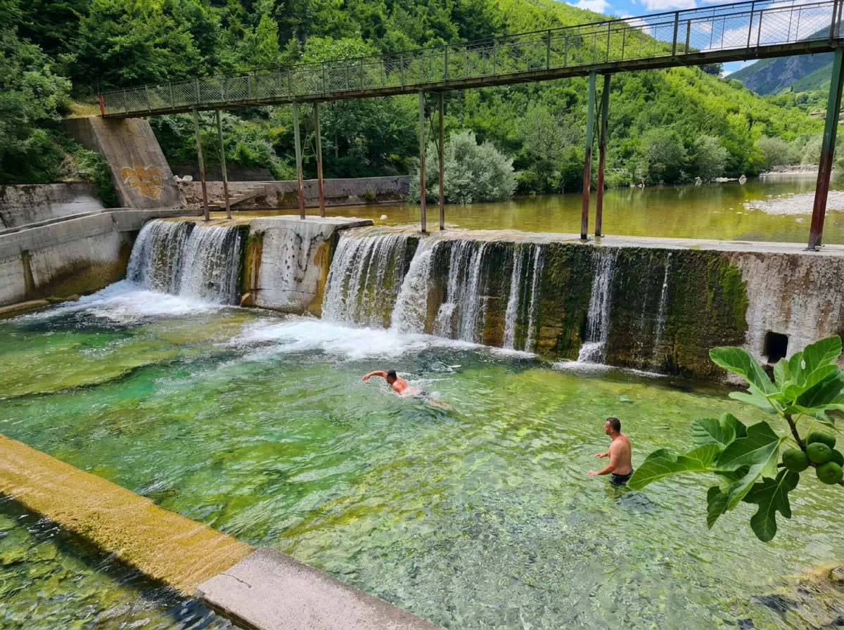 Badegäste, die im klaren, grünen Wasser der Drežanka vor einer Betonbarriere schwimmen, über die ein breiter, schäumender Wasserfall fließt, umgeben von Grün
