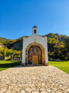 A stone path leading to the white facade of the monastery church in Dobrun, with a large, vivid fresco above the entrance door under the open sky