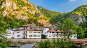 A view of the konaks and buildings of the Dobrun monastery complex, set at the foot of tall, steep cliffs and green hills