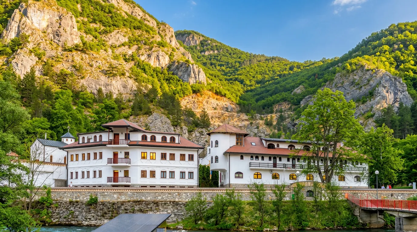 A view of the konaks and buildings of the Dobrun monastery complex, set at the foot of tall, steep cliffs and green hills