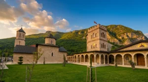 A view of the stone Church of Saint Nicholas of Myra and the tall defensive tower in the Rmanj monastery complex, surrounded by a well-kept green lawn and wooded hills.