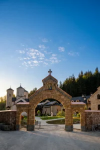 Monumentalna kamena lučna kapija na ulazu u manastirski kompleks Stuplje sa krstom na vrhu, kroz koju se u pozadini vide obrisi manastirske crkve i konaka pod plavim nebom