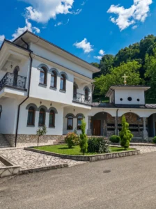 A view of the modern white monastery konak with arches, a paved courtyard, and landscaped green areas framed by dense forest in the background
