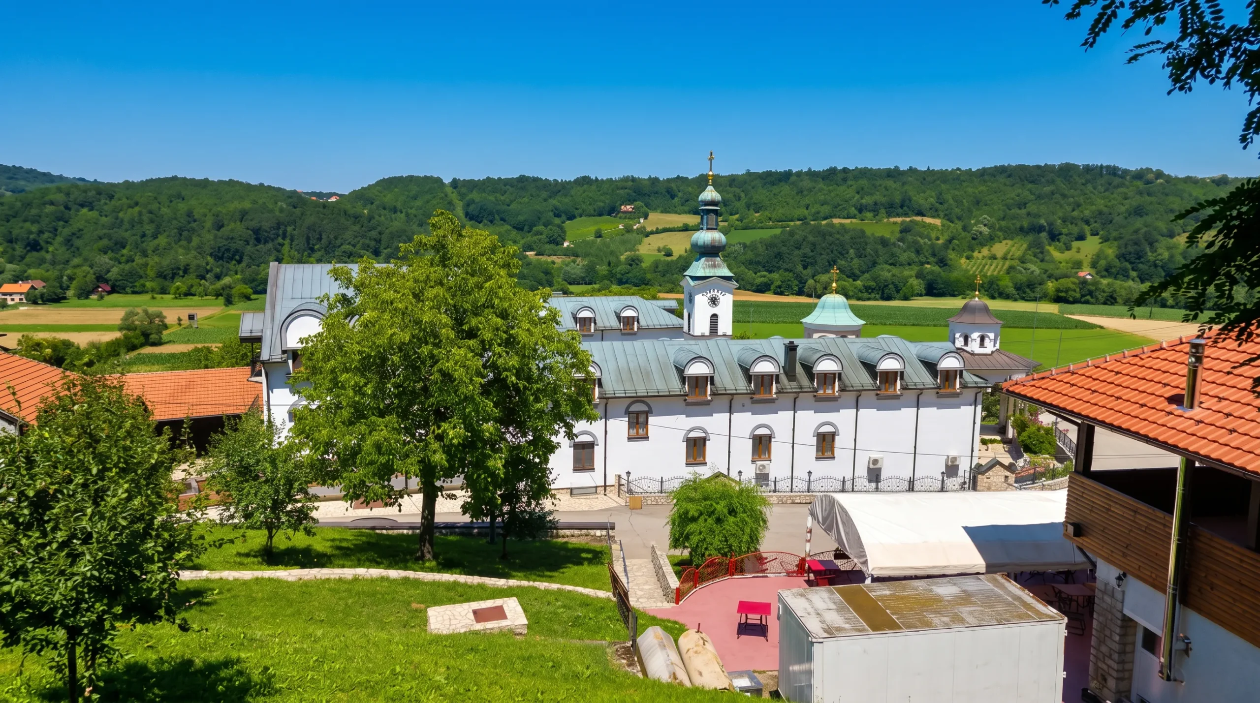 A view from a hill of the Tavna monastery complex, surrounded by green hills, fields, and dense forest under bright sunlight