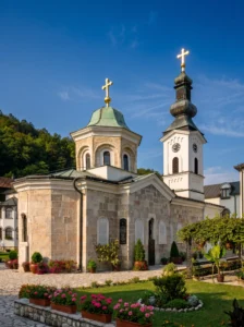 The stone church of Tavna Monastery with a dome and a Baroque bell tower decorated with golden crosses, set in a landscaped courtyard with flowers under a blue sky