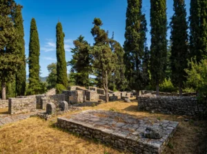 Remains of Roman stone walls and foundations of the historic site Mogorjelo, surrounded by dry grass and tall, slender cypress trees under a blue sky