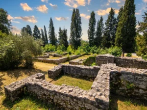 A close view of the regular shapes and partitions of ancient stone walls in Mogorjelo, with a row of tall cypress trees and olive trees in the background on a warm sunny day